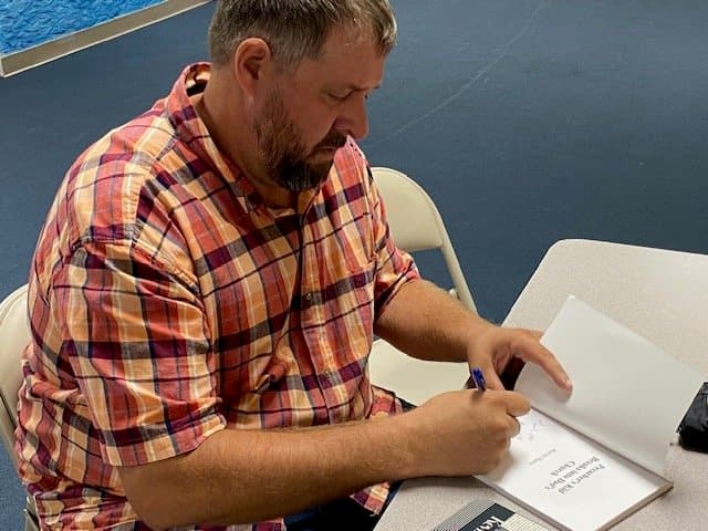 A man with a beard signing a copy of his book at a table.